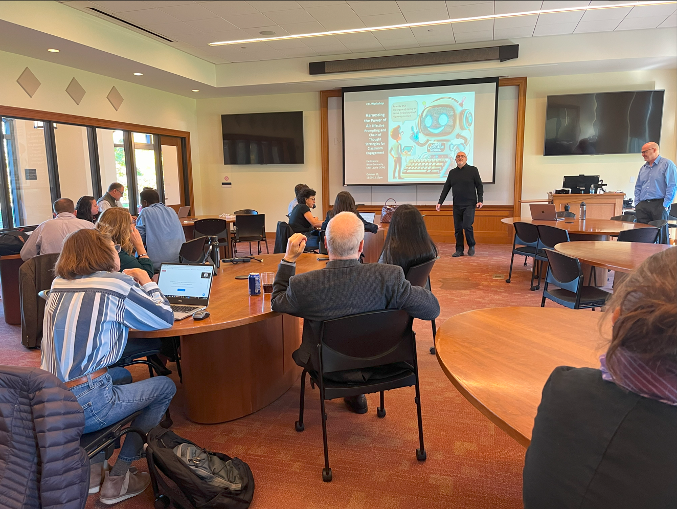 A classroom workshop with participants seated at round tables, listening to two presenters. A slide titled 