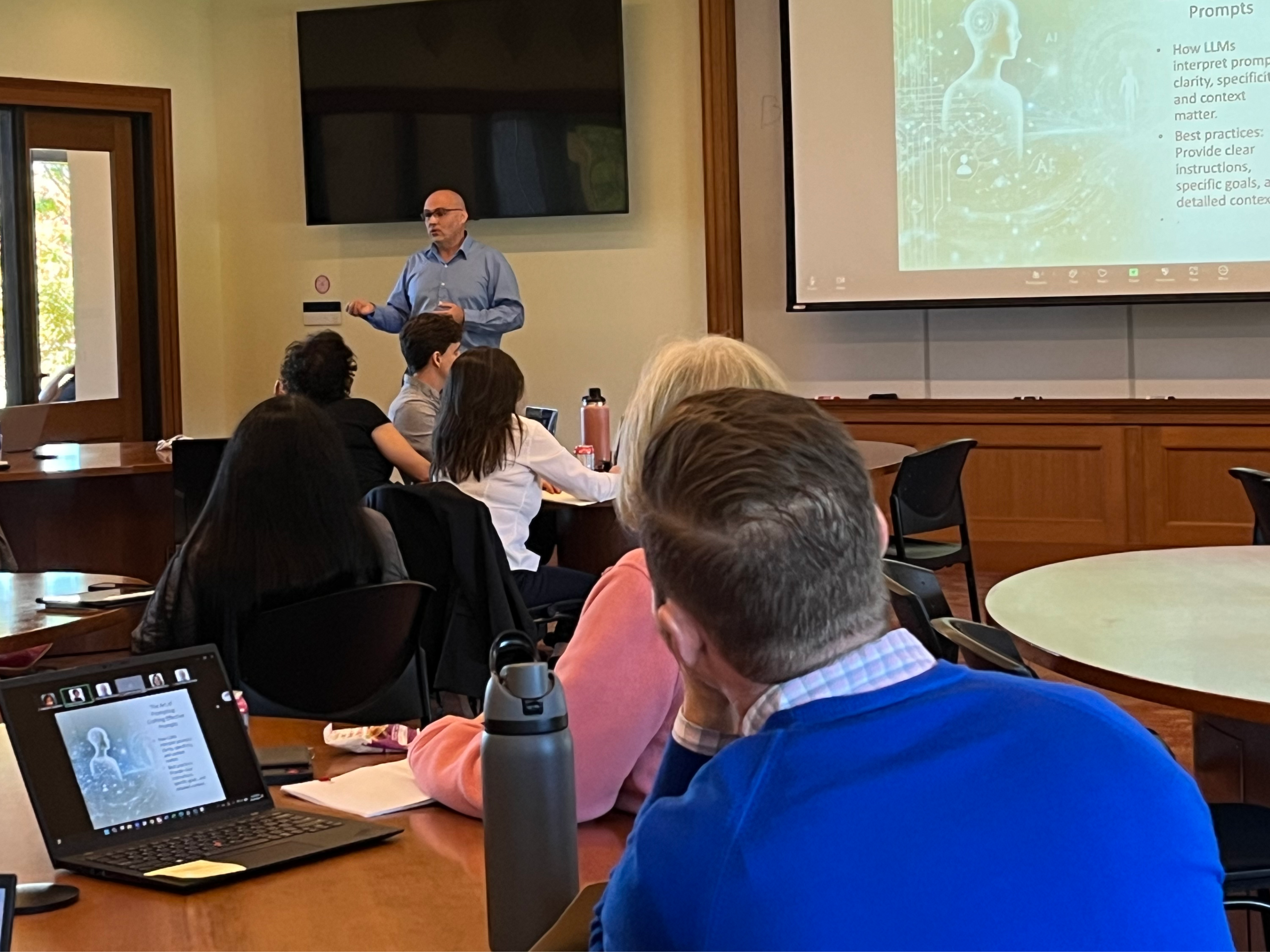 A speaker stands at the front of a classroom, presenting to an audience seated around tables. A projector screen shows a slide, while attendees take notes and engage.