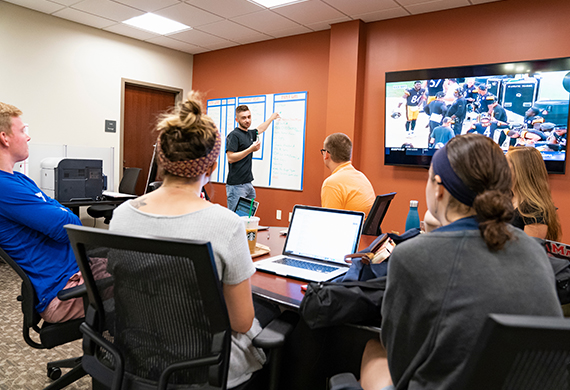 Students actively engage in a presentation around a conference table in a casual yet focused learning environment.