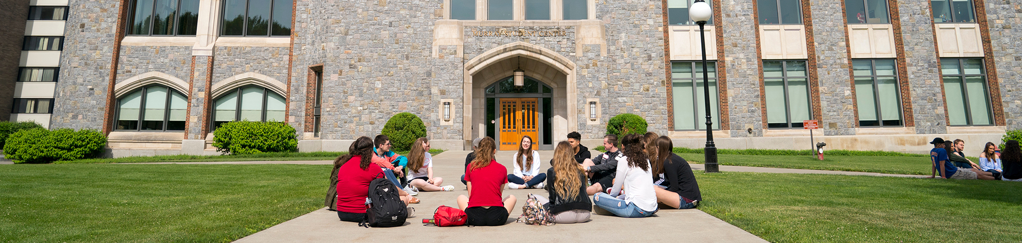 This is a background image of a group of students sits in a circle on a concrete path leading to a Marist Student center building with stone walls and arched doorways.