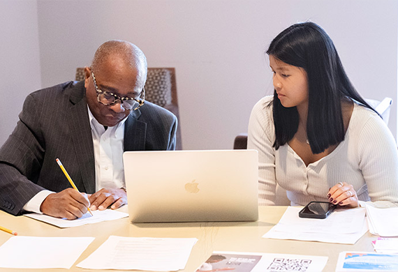 A faculty professor writes on paper with a pencil while a student sits beside him, with a laptop placed between them and papers and a phone nearby.