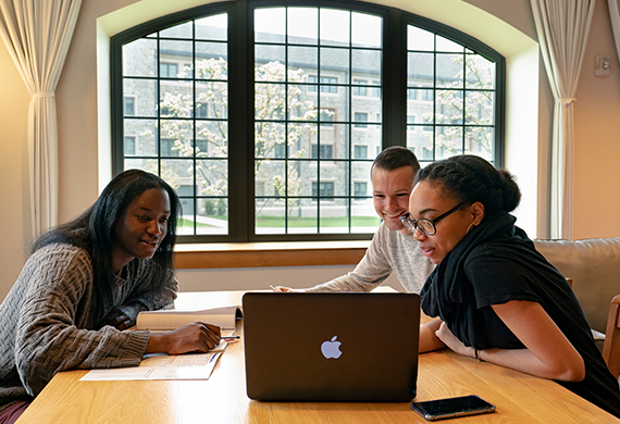 Three people are gathered around a laptop at a wooden table, likely engaged in a collaborative task or study session.