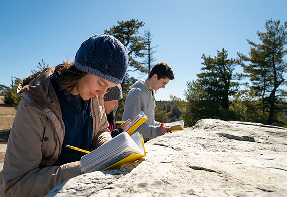 Image of Three people, One women and two men  study on a rock with a blue sky and pine trees behind them.