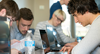 A group of young men collaborates in a casual workspace, using laptops and printed materials, conveying teamwork and focus.