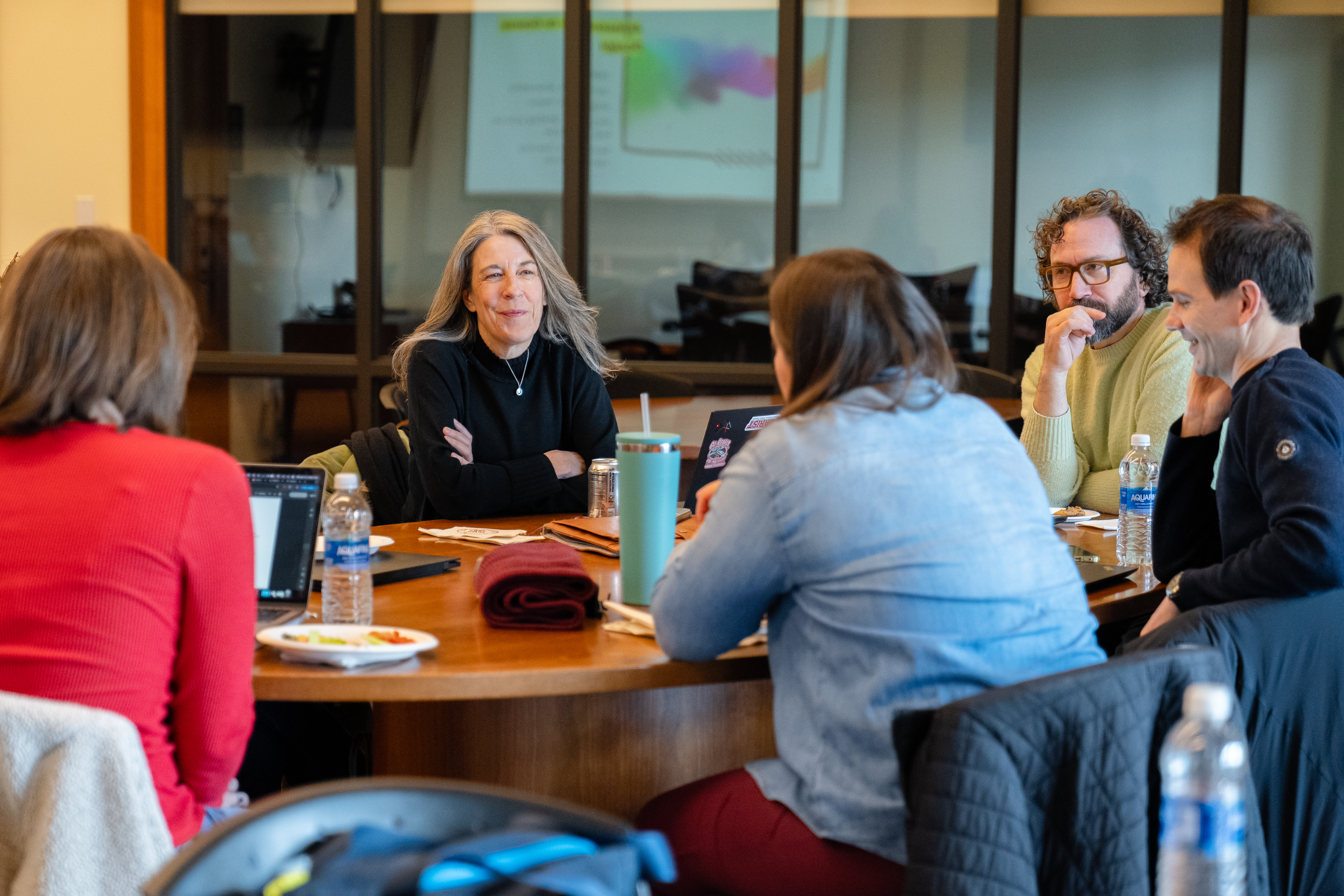 The image depicts a diverse group of five adults seated around a round wooden table in a well-lit office or meeting space, engaged in a relaxed yet focused conversation.