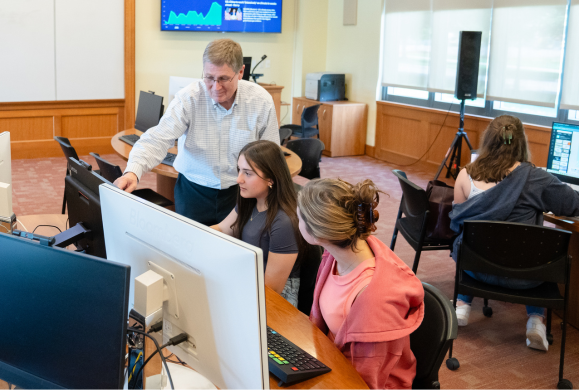 A teacher assists two students in a well-lit computer lab, with multiple workstations arranged around a central area, creating a cooperative and focused learning environment.