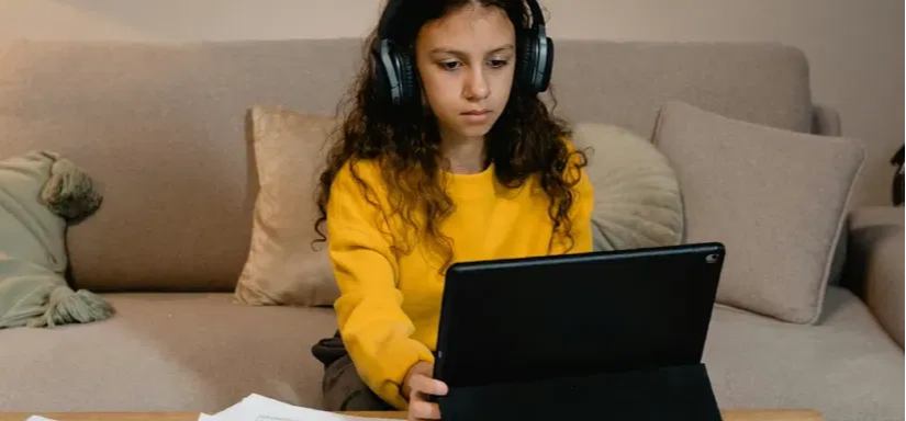 Image of a girl sitting on a couch wearing headphones and has a laptop in front of her. 